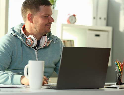 Mann mit blauem Kapuzenpullover und Kopfhörern um den Hals sitzt an einem Tisch mit Laptop, Kaffeetasse und Stifthalter in einem hellen Raum mit Bücherregal im Hintergrund