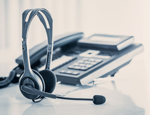Schwarzes Headset mit Mikrofon vor einem Tischtelefon auf hellem Tisch in unscharfem Hintergrund